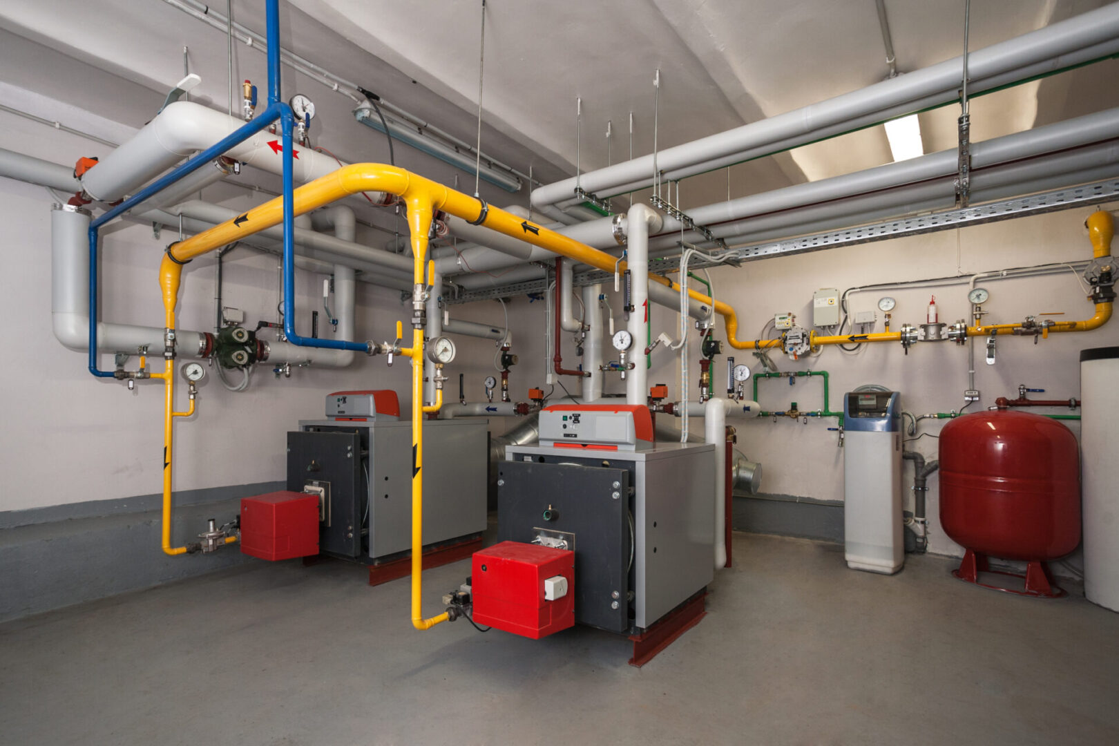 Room with industrial boilers, pipes in various colors, valves, gauges, and a water tank on the right. Clean and organized setup.