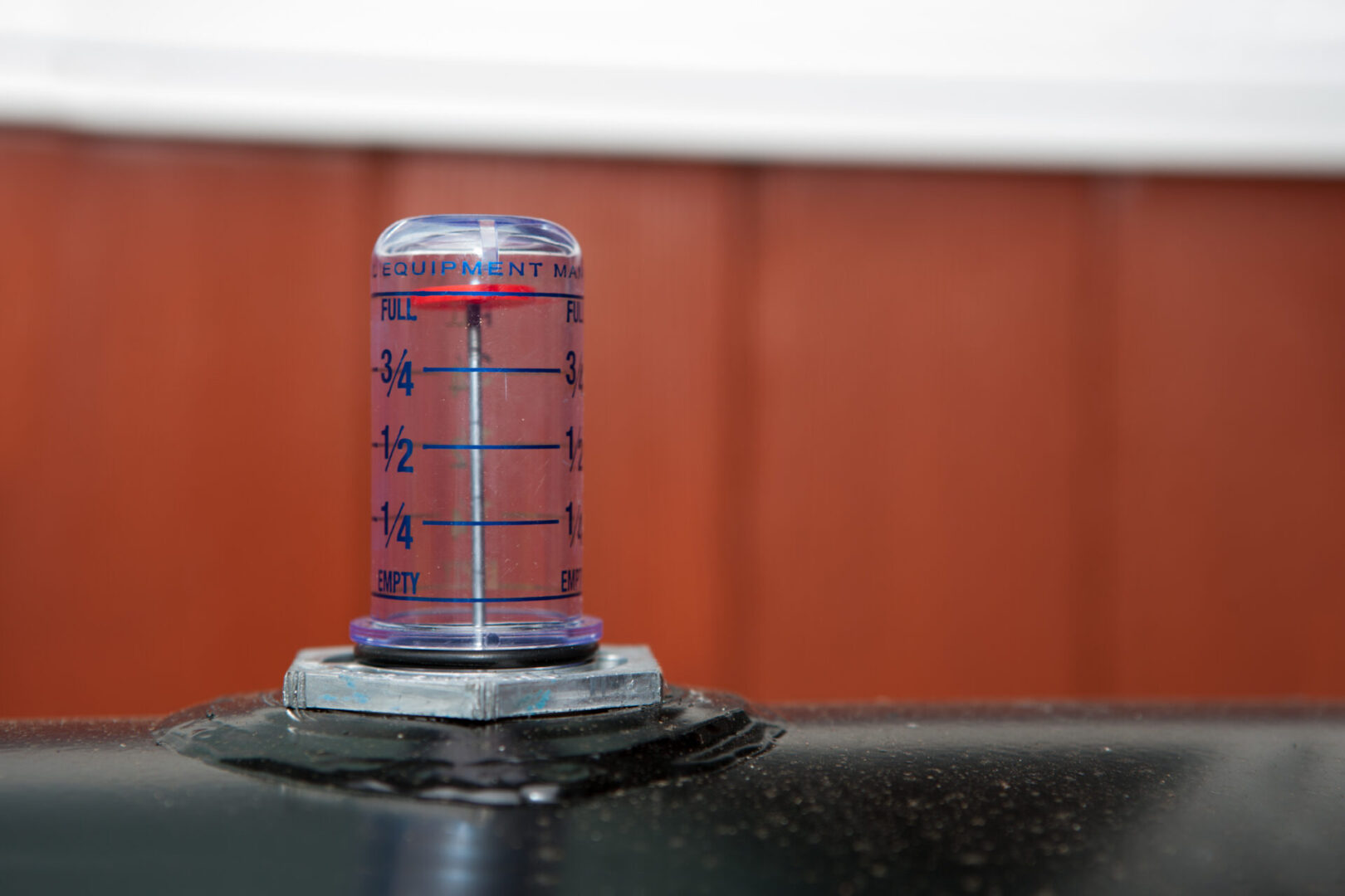 Close-up of a cylindrical gauge with clear plastic showing liquid levels marked from "Empty" to "Full." The gauge is attached to a black surface, with a blurred brown and white background.